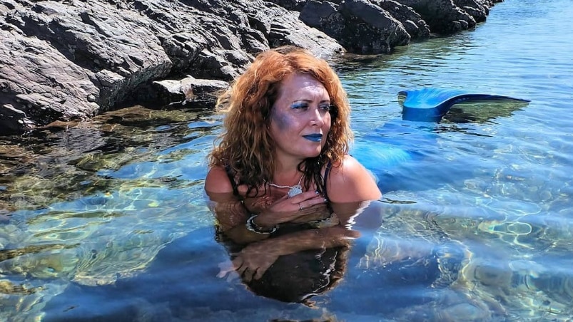 Nicola aka Naqulaan wears glitter makeup, resting on her elbows in a rock pool, with head shoulders and fins above water.