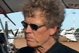 A TV still image of a man wearing sunglasses and a black polo shirt standing in front of a red and yellow gyrocopter.