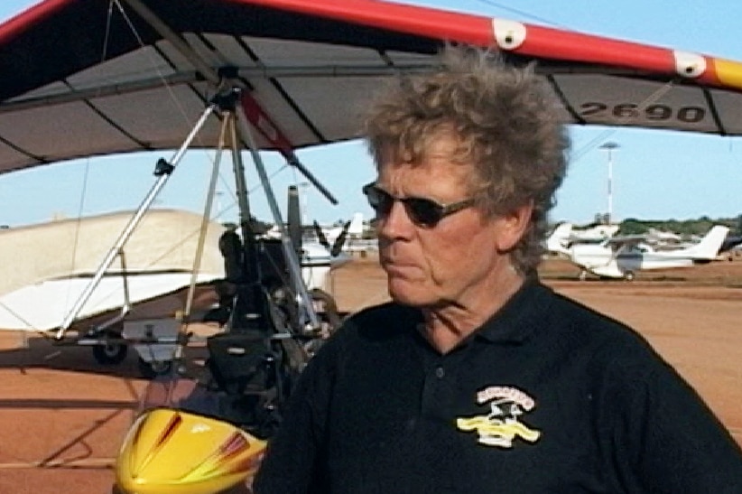 TV still image of a man wearing sunglasses and a black polo shirt standing in front of a red and yellow gyrocopter.