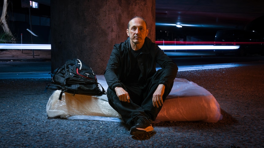 A man in black sits next to a backpack on a mattress on a city street