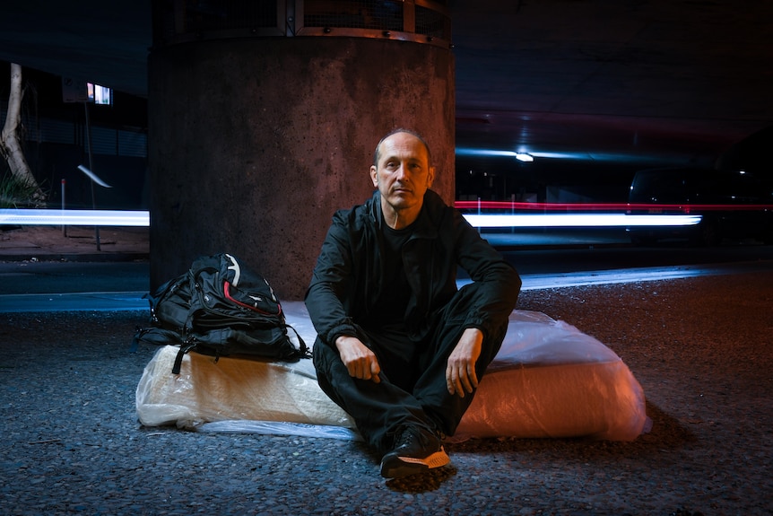 A man in black sits next to a backpack on a mattress on a city street