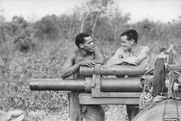 A black and white photo of two men without shirts leaning on a gun artillery.