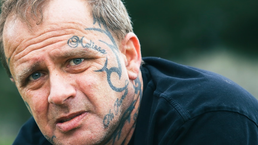 A close-up photograph of Brian Warton, who has face tattoos and piercing blue eyes.