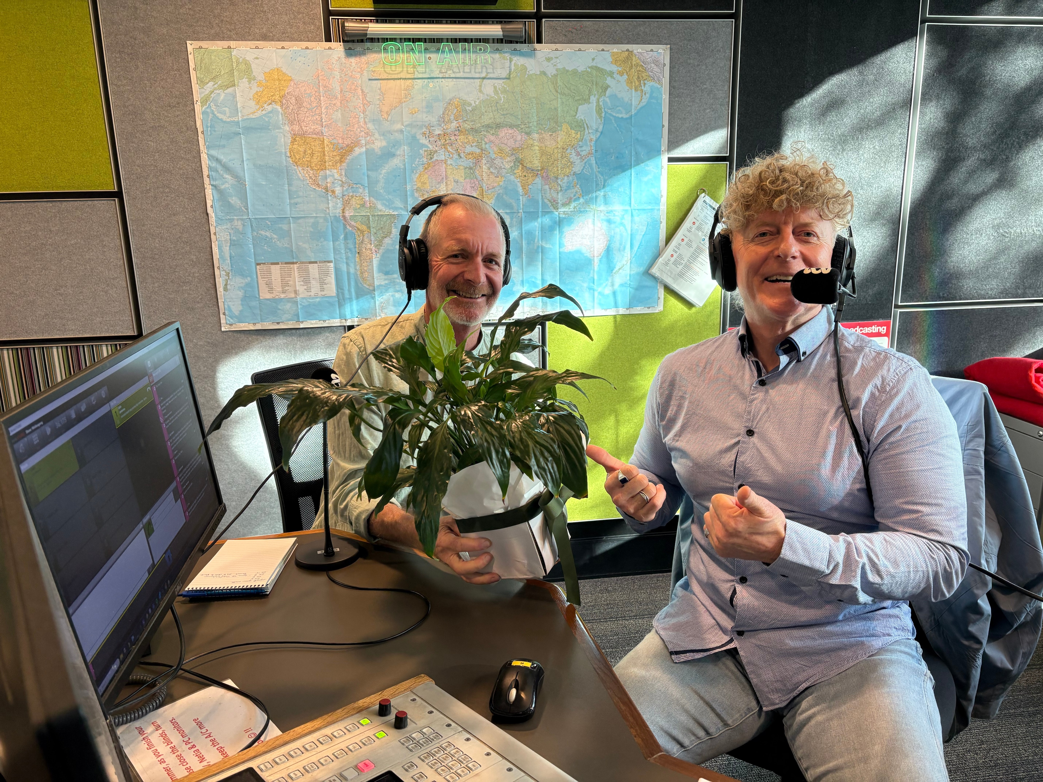 John and Mark wear microphone headsets in a studio and John holds a plant.