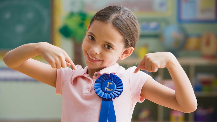A child points to a large blue 'first place' ribbon placed prominently on their chest.