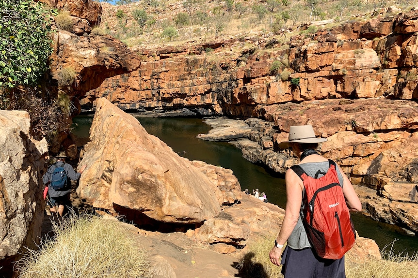 Tourists walk down a rocky path to a gorge, one woman wars hat, has red backpack.