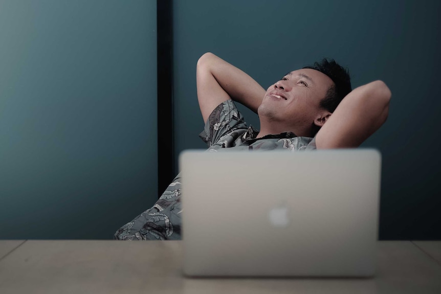 A man leans back on his chair, smiling, with an open laptop in front of him.