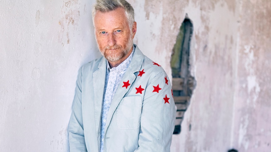 Billy Bragg has a slight smile and is wearing a light jacket with red stars on the left shoulder, in front of a broken wall