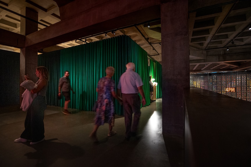 A dimly lit exhibition space with a dark green curtain, people walk around.