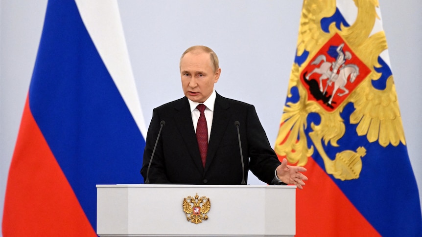Vladimir Putin speaks at podium with Russian flags behind him.