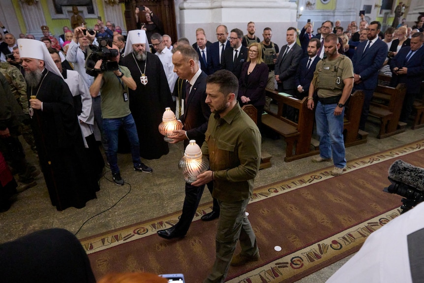 Zelenskyy wearing green walking down a church aisle holding a lantern next to another man