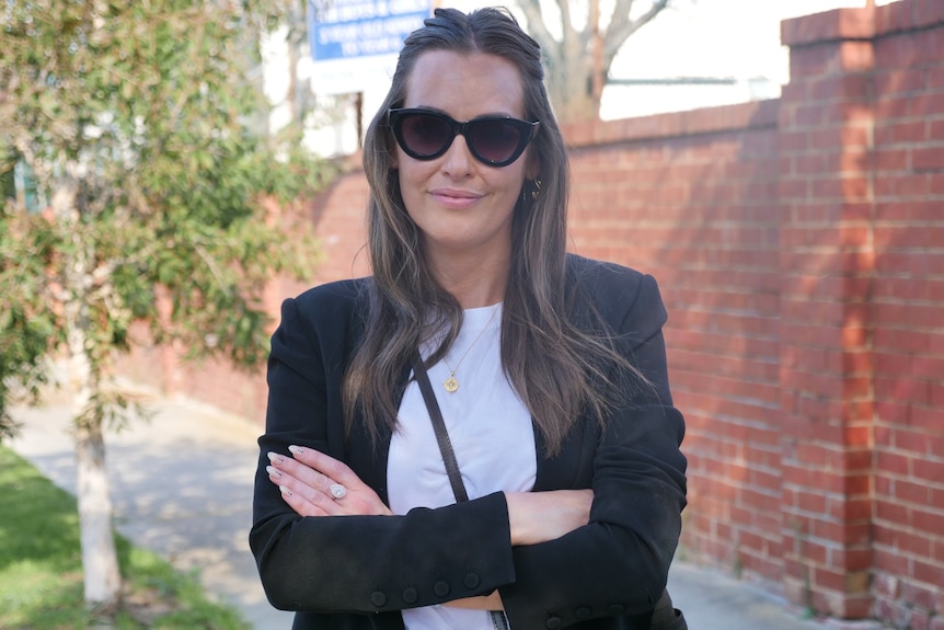 A woman with a black jacket and sunglasses folds her arms.