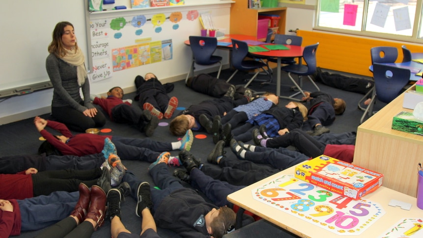 A teacher sits on the classroom floor with a group of children laying on the floor practising mindfulness.