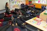 A teacher sits on the classroom floor with a group of children laying on the floor practising mindfulness.