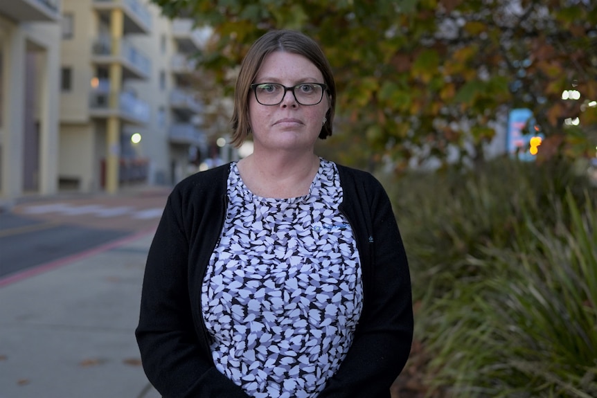 A woman with mid-length hair and wearing glasses looks into the camera with a serious expression. 