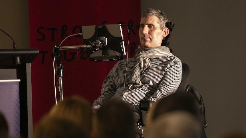 A man sits in a wheelchair and a small screen is positioned in front of his face.