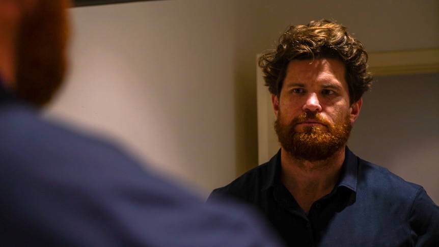 A man wearing a blue shirt stares at his reflection in a mirror