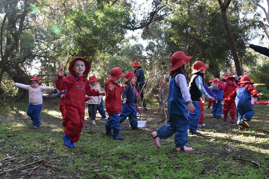 Children in red and blue jumpers and hats dance around a bush clearing.