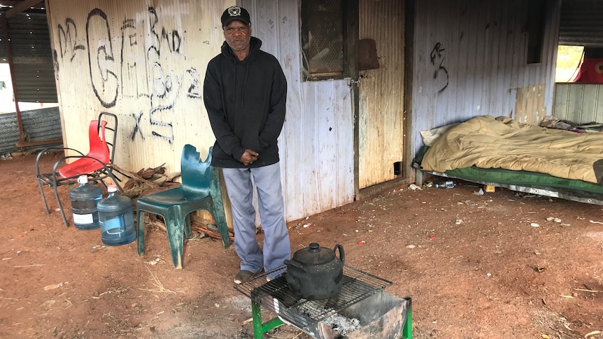 Dion Williams stands outside his tinshed home, with a teapot over the fire.