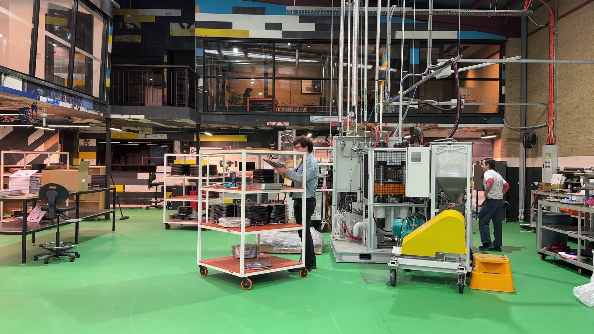 Dos hombres trabajan en una planta de prensado de discos con suelos verdes, grandes máquinas y carros.