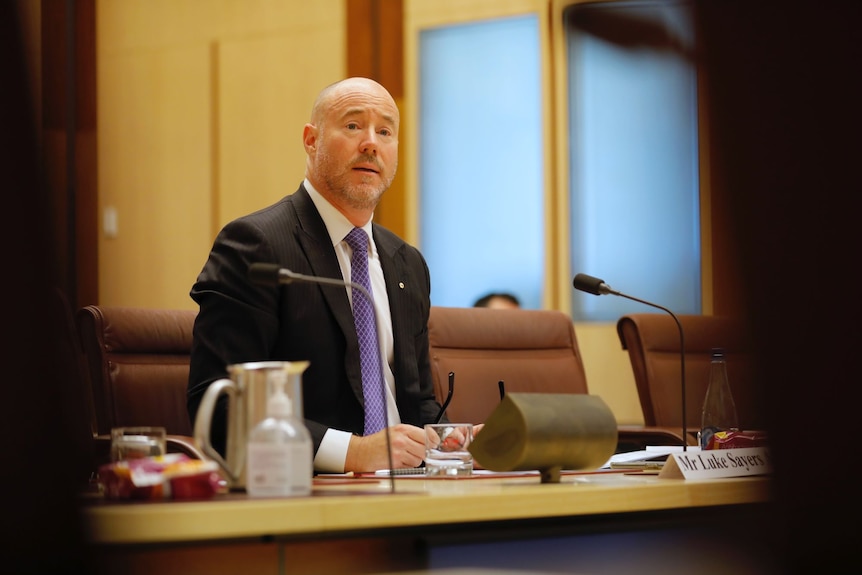 A bald man sits alone at a long table with a microphone. He is wearing a black suit, white collared shirt and purple tie.