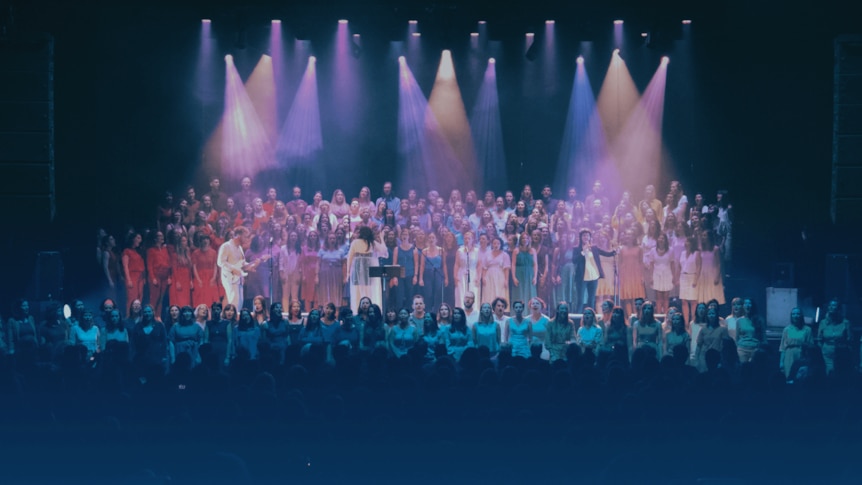 a large choir on stage with colourful lights beaming down on them.