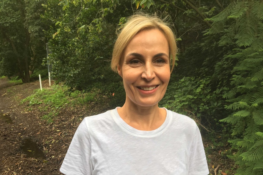 Penny Taylor, a woman with blonde hair, smiles at the camera in front of green trees.