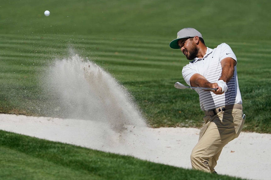Jason Day, wearing a white top and khaki trousers, plays a shot out of a bunker, spraying sand in front of him