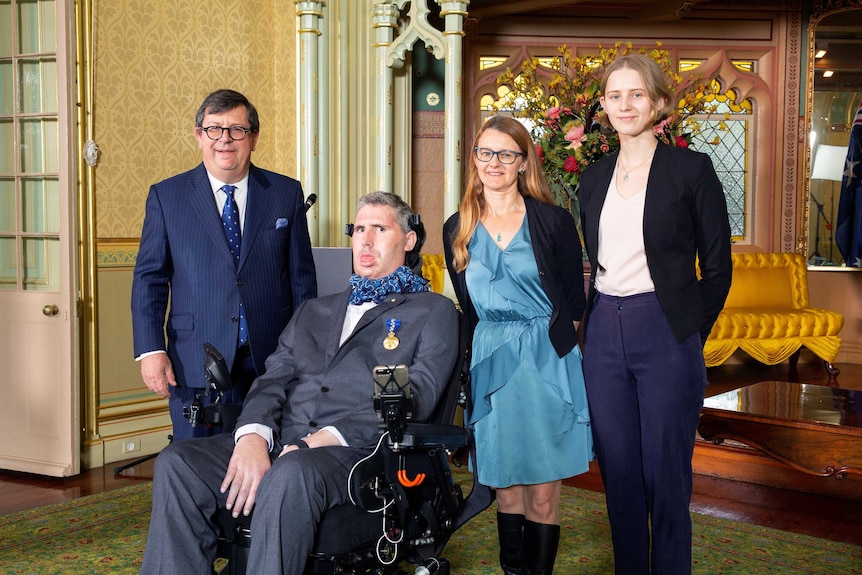 A man in a wheelchair, wearing a suit, is flanked by two women and a man in a dark suit.