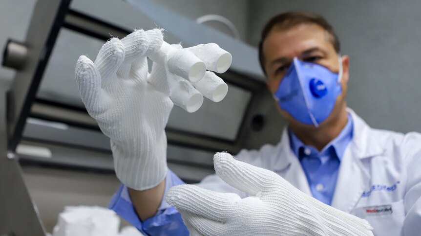 Man in white coat and blue mask holds up  small set of white tubes wearing thick white gloves in a lab type setting