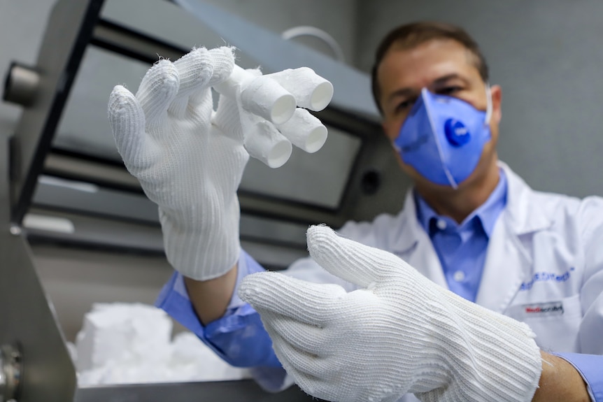 Man in white coat and blue mask holds up  small set of white tubes wearing thick white gloves in a lab type setting