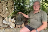 An older man sits in a garden next to an old bronze pearl divers helmet