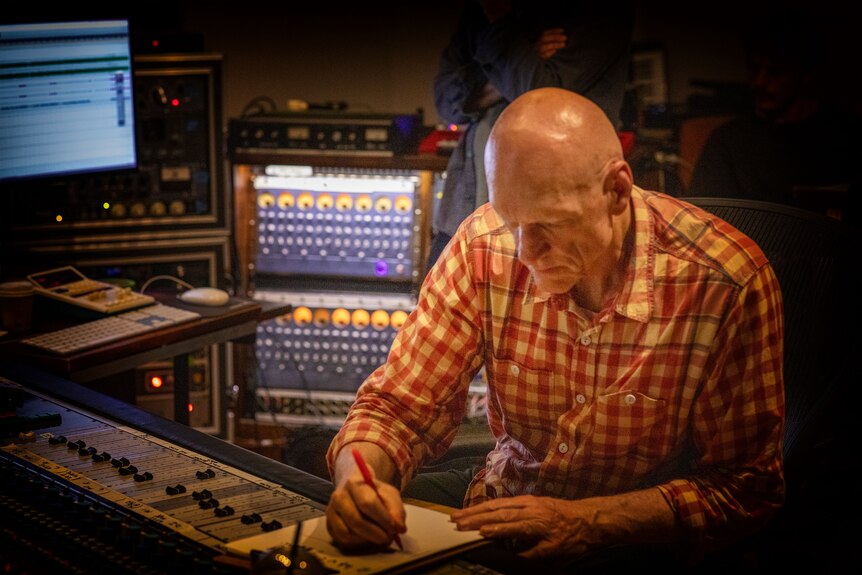 Peter Garrett sits in music studio seated at table writing in a notebook, his head down