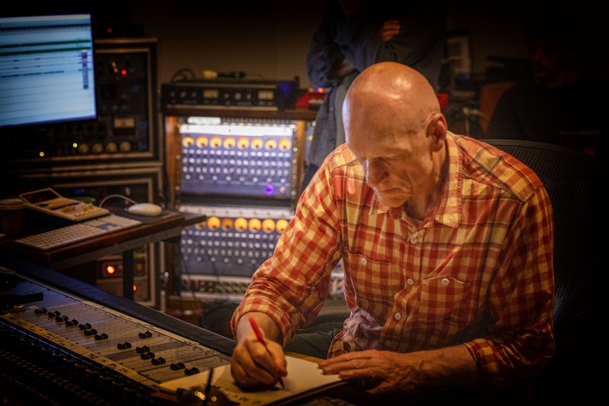 Peter Garrett sits in music studio seated at table writing in a notebook, his head down