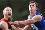 A Melbourne AFL players pushes against a North Melbourne opponent as they look to the sky waiting for the ball to be thrown in.