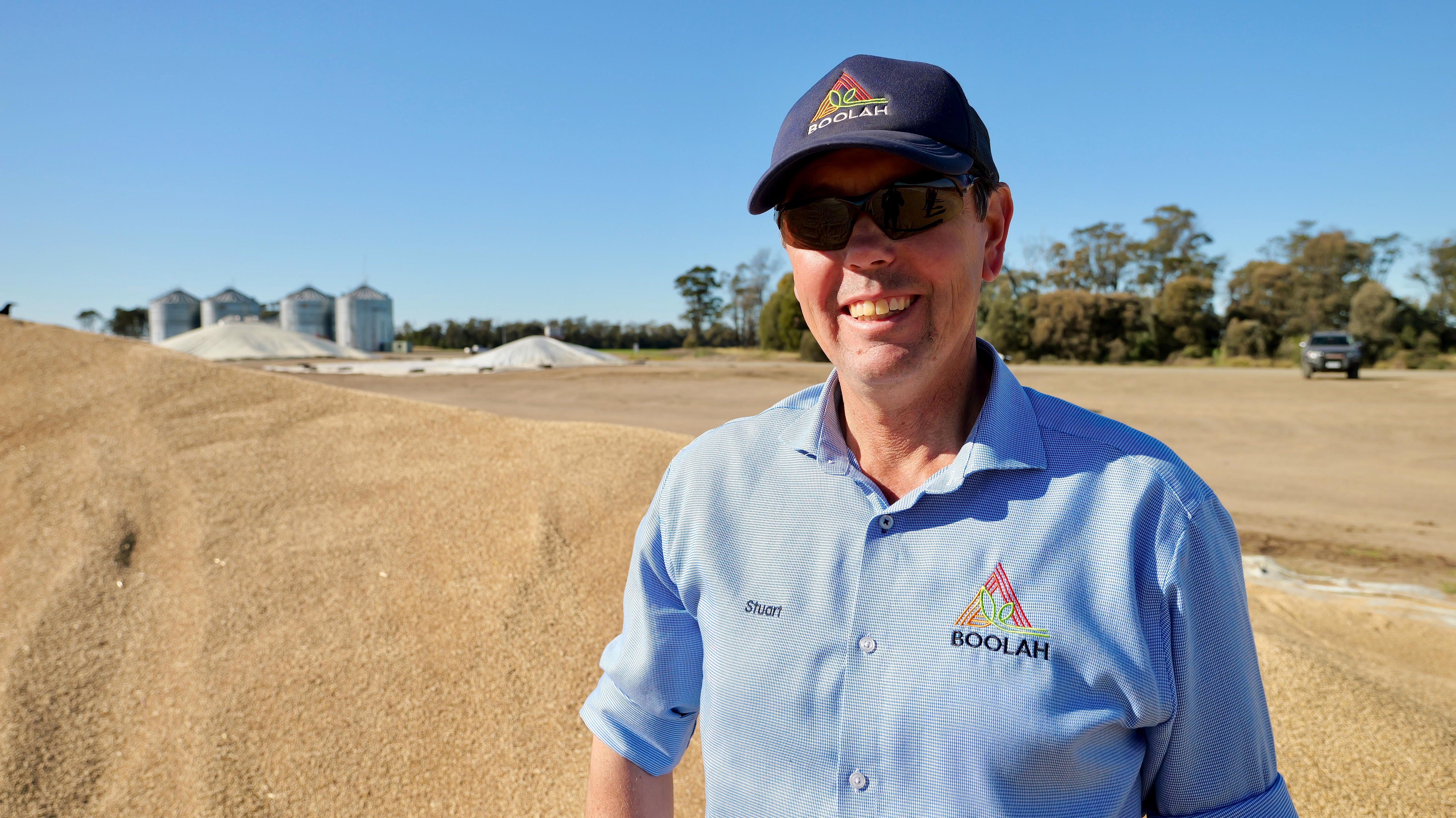 A man in a navy cap and a chambray blue shirt stands smiling in front of a grain pile and silos in the distance.