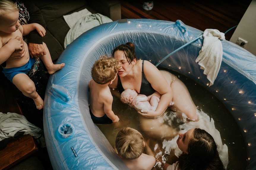 A woman cradles a newborn baby in a birthing pool while talking to other young children.