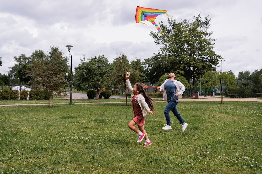 A young girl runs and holds a rainbow kite smiling while her dad laughs and runs behind her in a park.