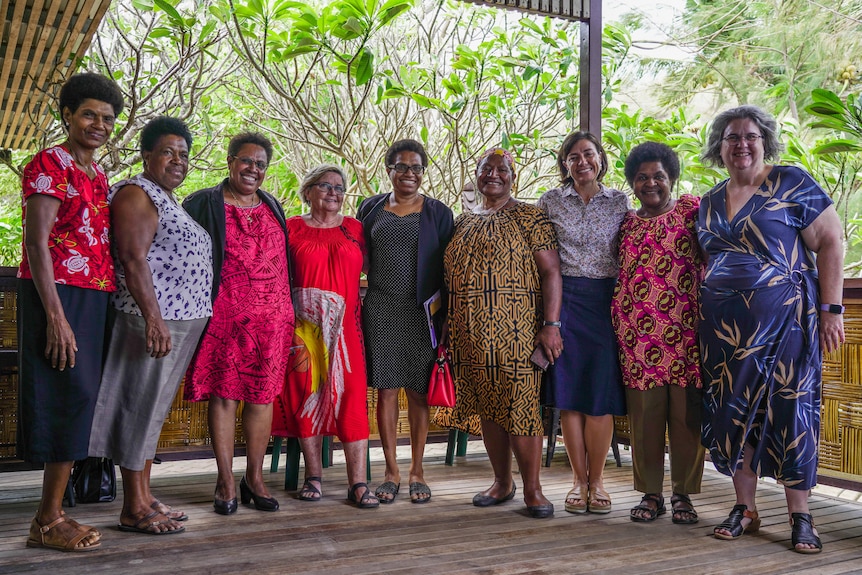 Nine women dressed in different brightly coloured attire stand side-by-side against a tropical backdrop