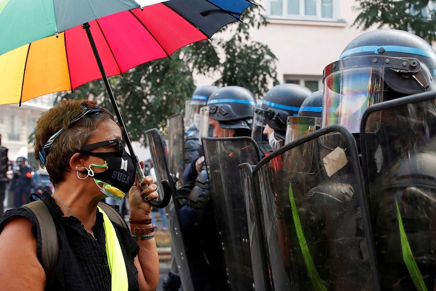 Woman wearing mask, holds rainbow umbrella, stands in front of riot police with shields