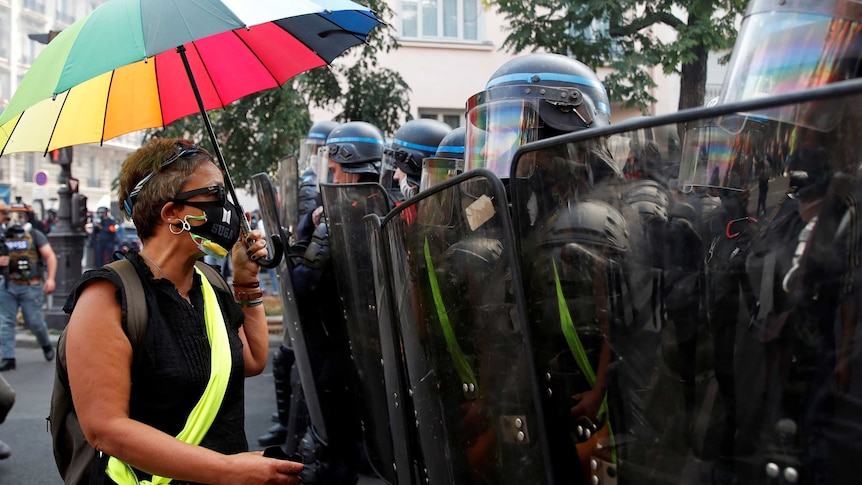 Woman wearing mask, holds rainbow umbrella, stands in front of riot police with shields