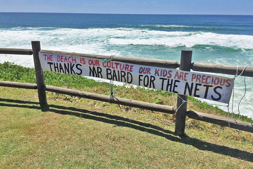A sign that reads "Thanks Mr Baird for the nets" hanging on a wooden fence in front of the ocean.
