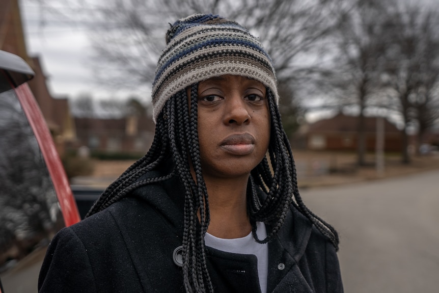 A close up of a Black woman with dreadlocks and wearing a rainbow beanie and black jacket frowns.