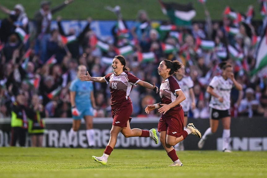 Two women soccer players wearing maroon and white celebrate during a game with Palestine flags in the background
