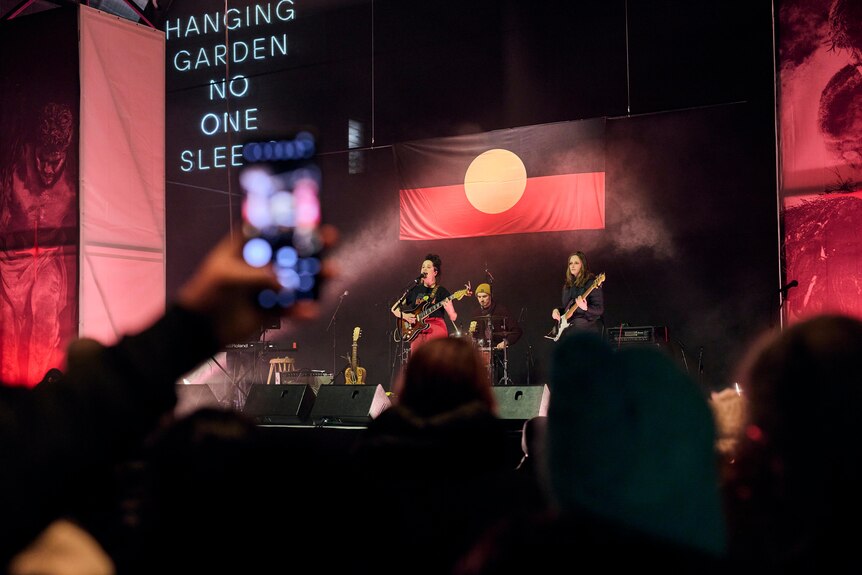 Band of three women perform onstage in front of an Aboriginal flag. A crowd member's phone can be seen in the foreground.