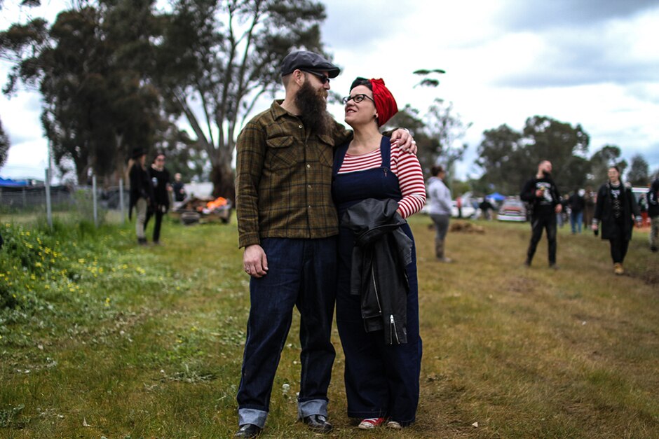 A man and a woman stand side by side, looking at each other. His arm is around her shoulders.