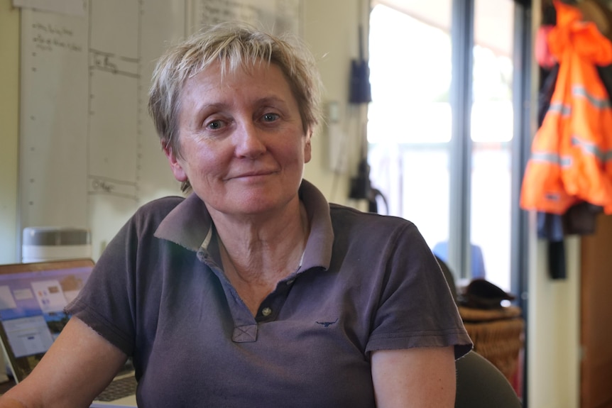 Woman with short blonde hair sitting in front of a workspace with desk and whiteboard.