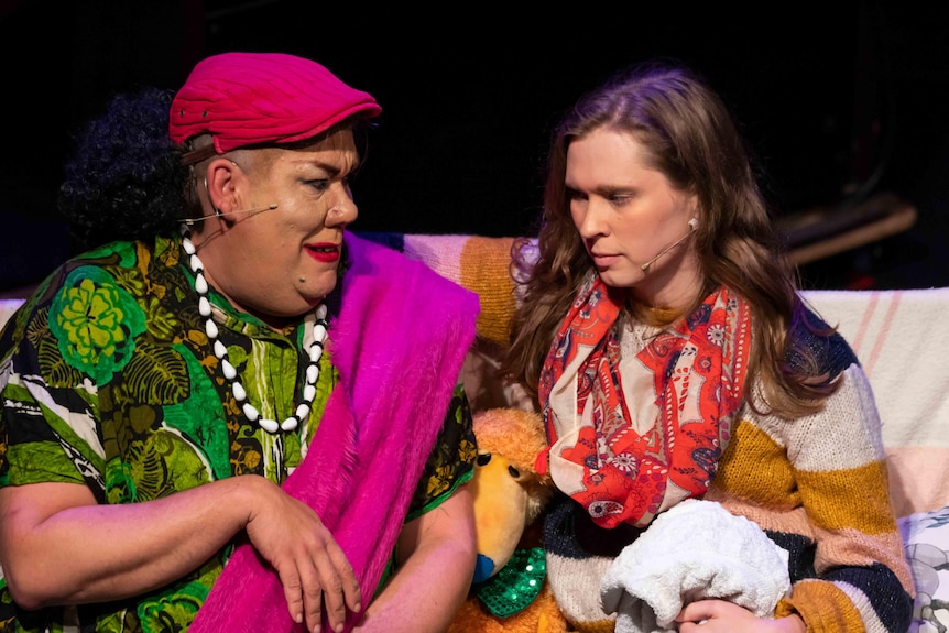 A colourful drag queen sits on a couch talking to a woman wearing a bright scarf.