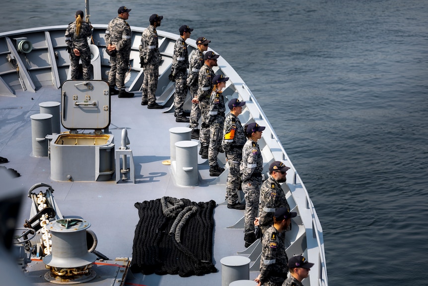 A dozen men in military uniforms stand around the rim of a ship floating on water