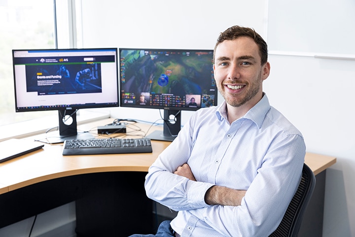 A man in a light-blue shirt sits in front of two computer screens.